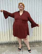 Load image into Gallery viewer, Additional full-body front view of size 3 Torrid brick red plaid pattern bell sleeve button-up shirt dress styled with black heels on a size 22/24 model. The photo is taken outside in natural lighting.
