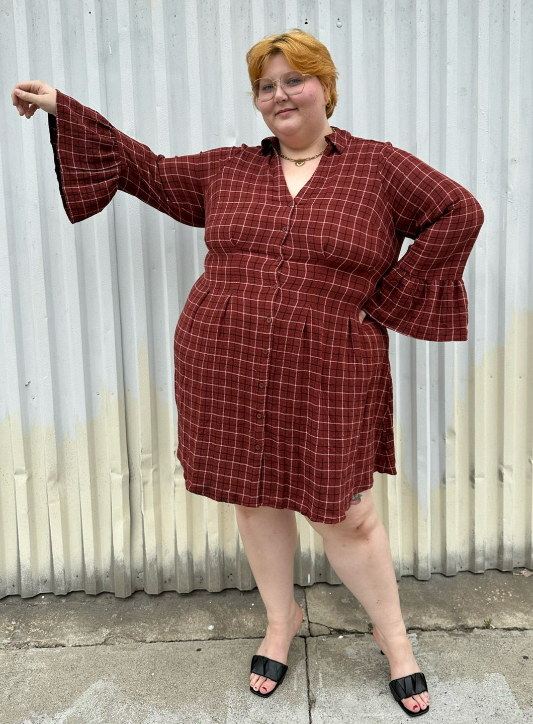 Full-body front view of size 3 Torrid brick red plaid pattern bell sleeve button-up shirt dress styled with black heels on a size 22/24 model. The photo is taken outside in natural lighting.