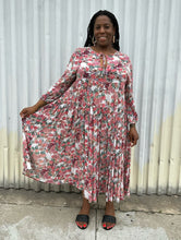Load image into Gallery viewer, Full-body front view of a size L RAGA red, green, and white rose patterned pleated maxi dress styled with black slides on a size 14/16 model. The photo is taken outside in natural lighting.

