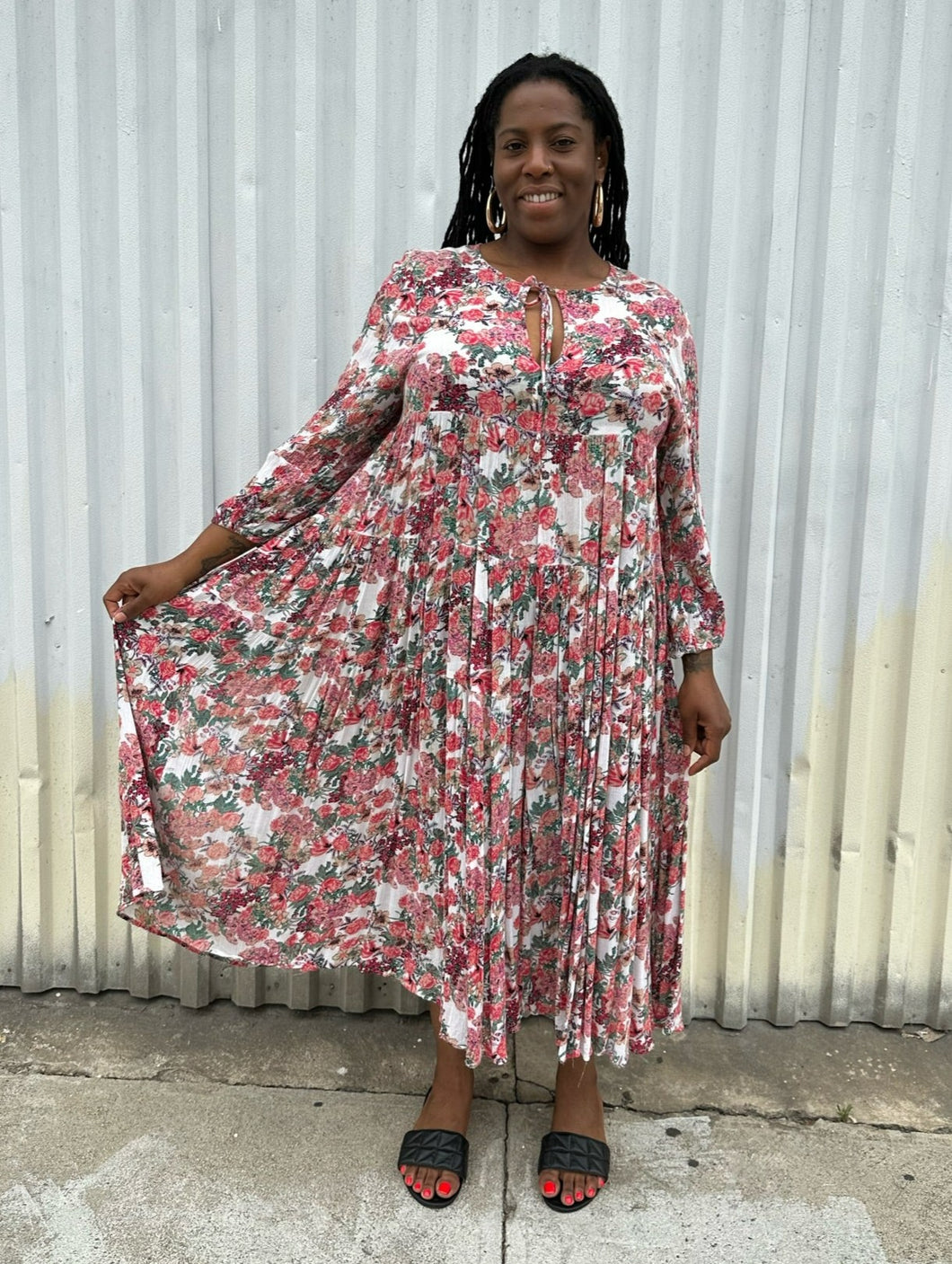 Full-body front view of a size L RAGA red, green, and white rose patterned pleated maxi dress styled with black slides on a size 14/16 model. The photo is taken outside in natural lighting.