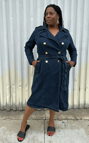 Full-body front view of a size 14/16 Eloquii dark wash denim trench coat dress with light brown buttons and tie belt styled with black slides on a size 14/16 model. The photo is taken outside in natural lighting.