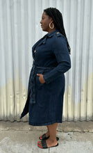 Load image into Gallery viewer, Full-body side view of a size 14/16 Eloquii dark wash denim trench coat dress with light brown buttons and tie belt styled with black slides on a size 14/16 model. The photo is taken outside in natural lighting.
