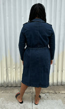 Load image into Gallery viewer, Full-body back view of a size 14/16 Eloquii dark wash denim trench coat dress with light brown buttons and tie belt styled with black slides on a size 14/16 model. The photo is taken outside in natural lighting.
