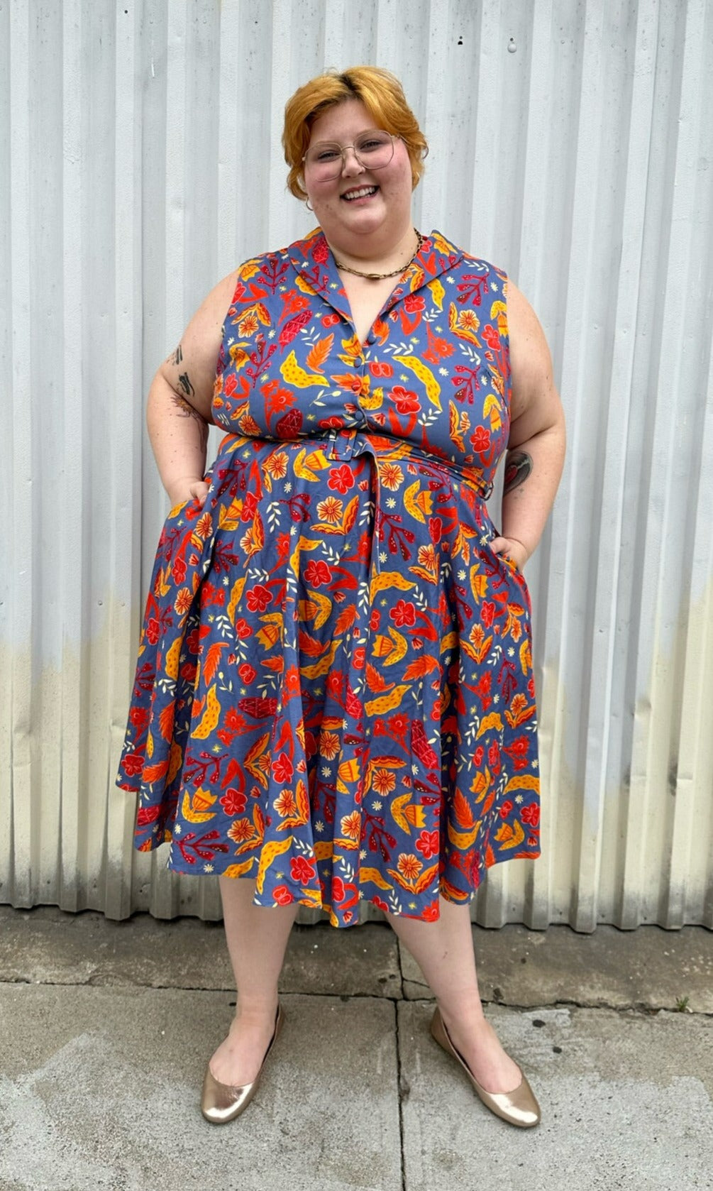 Full-body front view of a size 3X Miss Lulo blue, yellow, and red mixed floral print collared midi dress with fabric buttons and a matching belt styled with gold flats on a size 22/24 model. The photo is taken outside in natural lighting.