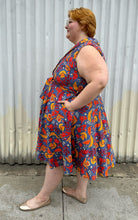 Load image into Gallery viewer, Full-body side view of a size 3X Miss Lulo blue, yellow, and red mixed floral print collared midi dress with fabric buttons and a matching belt styled with gold flats on a size 22/24 model. The photo is taken outside in natural lighting.
