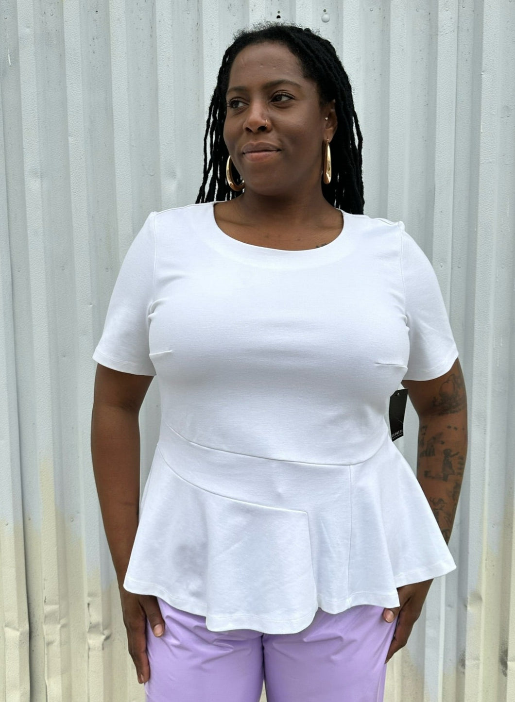 Front view of a size 16 Eloquii white tee with ruffle hem and asymmetrical stitching detail styled with lavender pleather pants on a size 14/16 model. The photo is taken outside in natural lighting.