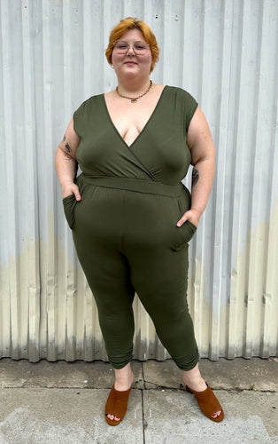Full-body front view of a size 3X Lillian olive green v-neck stretchy jumpsuit with pockets styled with brown mules on a size 22/24 model. The photo is taken outside in natural lighting.
