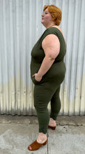 Load image into Gallery viewer, Full-body side view of a size 3X Lillian olive green v-neck stretchy jumpsuit with pockets styled with brown mules on a size 22/24 model. The photo is taken outside in natural lighting.
