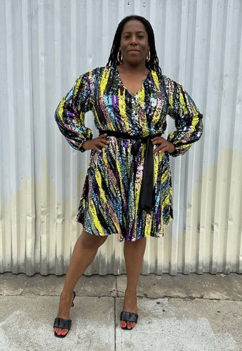 Full-body front view of a size 16 Eloquii yellow, blue, purple, pink and green sequin striped wrap dress with black belt styled with black heels on a size 14/16 model. The photo is taken outside in natural lighting.