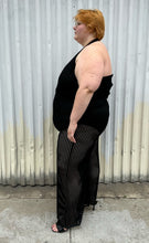 Load image into Gallery viewer, Full-body side view of a size 3 Fashion to Figure black halter sheer mesh vertical striped jumpsuit with a bodysuit lining styled with black heels on a size 22/24 model. The photo is taken outside in natural lighting.
