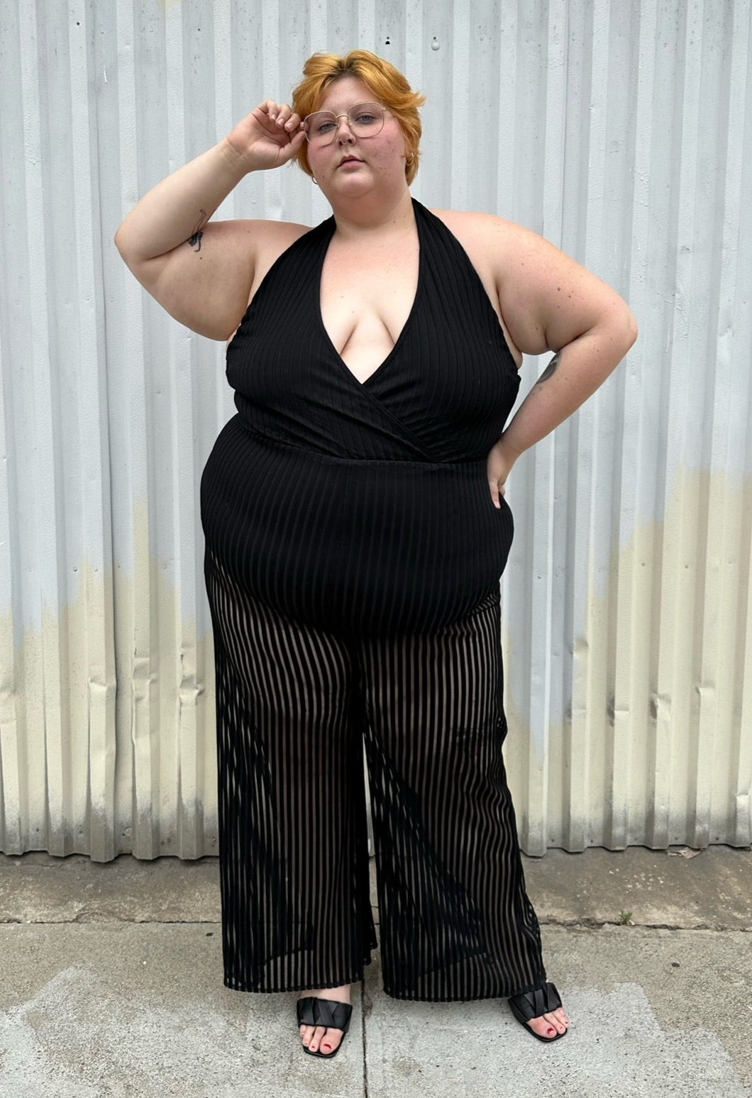 Full-body front view of a size 3 Fashion to Figure black halter sheer mesh vertical striped jumpsuit with a bodysuit lining styled with black heels on a size 22/24 model. The photo is taken outside in natural lighting.