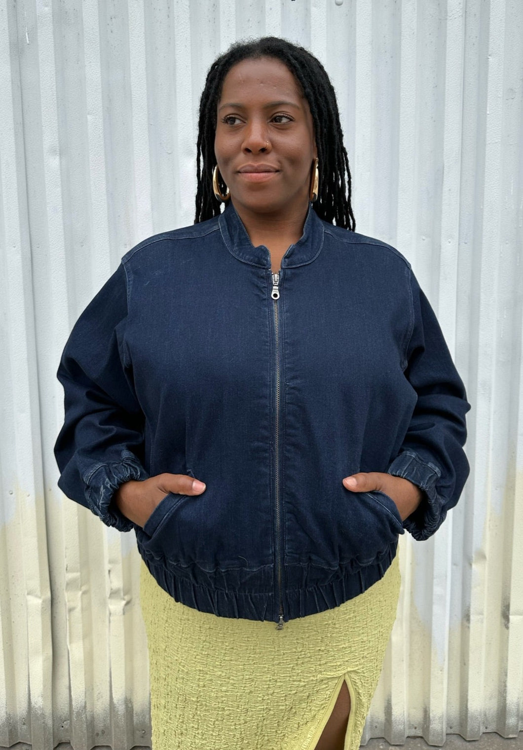 Front view of a size 14/16 Universal Standard dark wash denim bomber jacket styled zipped up over a chartreuse dress on a size 14/16 model. The photo is taken outside in natural lighting.