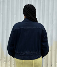 Load image into Gallery viewer, Back view of a size 14/16 Universal Standard dark wash denim bomber jacket styled zipped up over a chartreuse dress on a size 14/16 model. The photo is taken outside in natural lighting.
