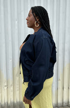 Load image into Gallery viewer, Side view of a size 14/16 Universal Standard dark wash denim bomber jacket styled open over a chartreuse dress on a size 14/16 model. The photo is taken outside in natural lighting.
