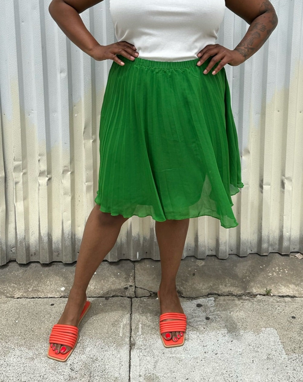 Front view of a size XL Elle Magazine kelly green pleated midi skirt styled with a white tank and orange slides on a size 14/16 model. The photo is taken outside in natural lighting.