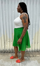 Load image into Gallery viewer, Full-body side view of a size XL Elle Magazine kelly green pleated midi skirt styled with a white tank and orange slides on a size 14/16 model. The photo is taken outside in natural lighting.
