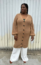 Load image into Gallery viewer, Full-body front view of a size 14/16 Eloquii light brown knit longline cardigan with subtle puff sleeves styled closed over white pants on a size 14/16 model. The photo is taken outside in natural lighting.

