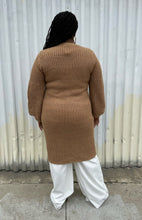 Load image into Gallery viewer, Full-body back view of a size 14/16 Eloquii light brown knit longline cardigan with subtle puff sleeves styled over white pants on a size 14/16 model. The photo is taken outside in natural lighting.

