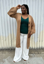Load image into Gallery viewer, Full-body front view of a size 14/16 Eloquii light brown knit longline cardigan with subtle puff sleeves styled open over a green top and white pants on a size 14/16 model. The photo is taken outside in natural lighting.
