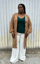 Load image into Gallery viewer, Additional full-body front view of a size 14/16 Eloquii light brown knit longline cardigan with subtle puff sleeves styled open over a green top and white pants on a size 14/16 model. The photo is taken outside in natural lighting.
