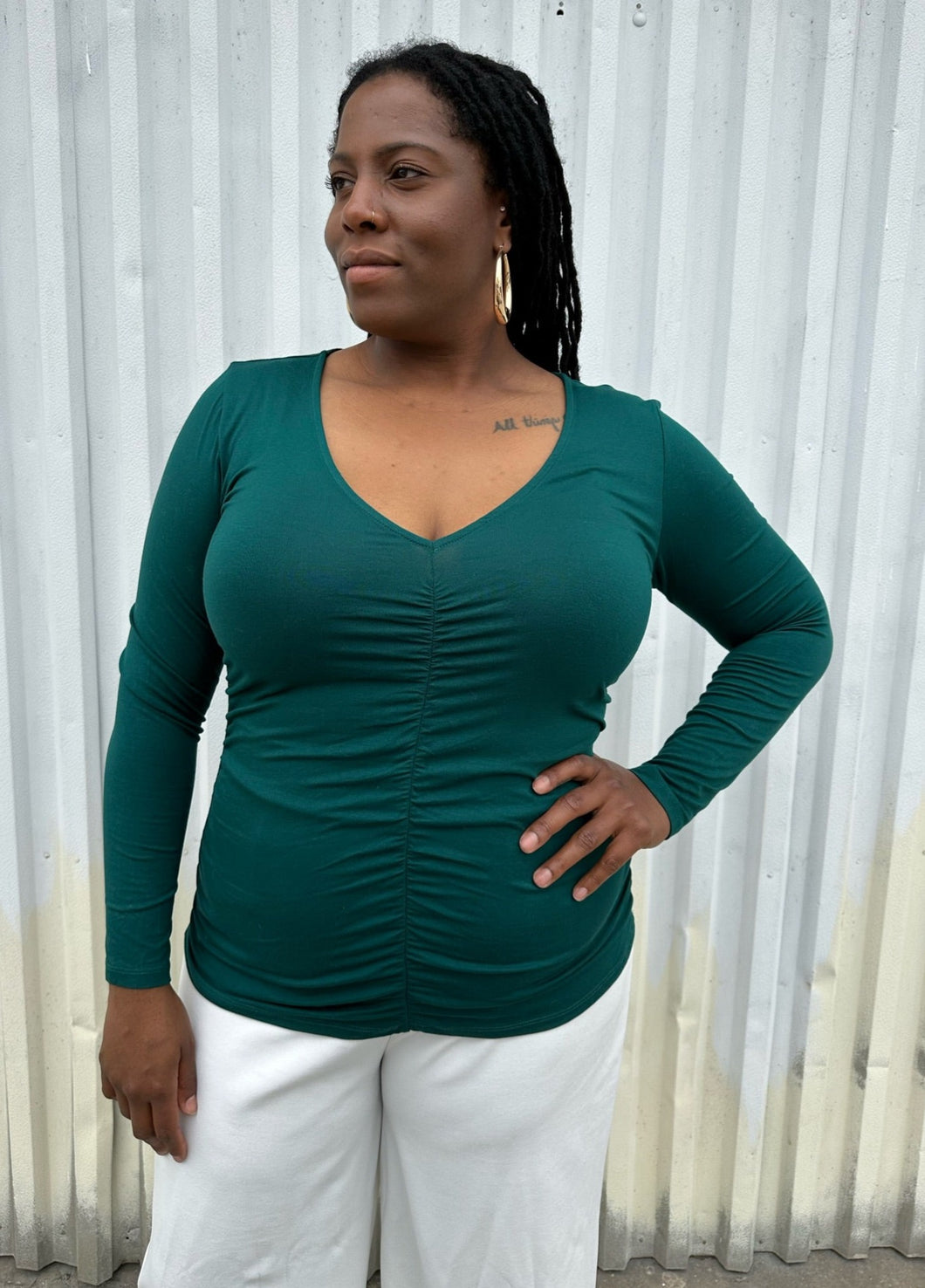 Front view of a size 14/16 ELoquii dark teal ruched front long sleeve tee styled with white pants on a size 14/16 model. The photo is taken outside in natural lighting.