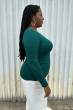 Load image into Gallery viewer, Side view of a size 14/16 ELoquii dark teal ruched front long sleeve tee styled with white pants on a size 14/16 model. The photo is taken outside in natural lighting.
