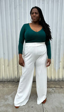Load image into Gallery viewer, Full-body front view of a size 14/16 ELoquii dark teal ruched front long sleeve tee styled tucked into white pants on a size 14/16 model. The photo is taken outside in natural lighting.
