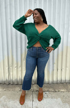 Load image into Gallery viewer, Full-body front view of a size 14/16 Eloquii emerald green twist-front cropped sweater with tummy cut-out styled with medium wash denim on a size 14/16 model. The photo is taken outside in natural lighting.
