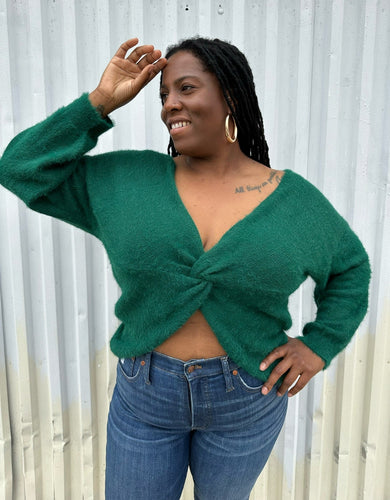 Front view of a size 14/16 Eloquii emerald green twist-front cropped sweater with tummy cut-out styled with medium wash denim on a size 14/16 model. The photo is taken outside in natural lighting.