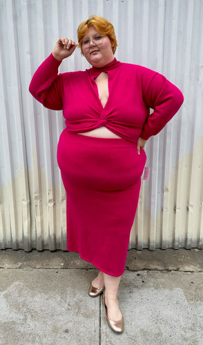 Full-body front view of a size 22/24 Eloquii hot pink sweater dress with turtleneck top, keyhole bust, and belly cut-out styled with gold flats on a size 22/24 model. The photo is taken outside in natural lighting.