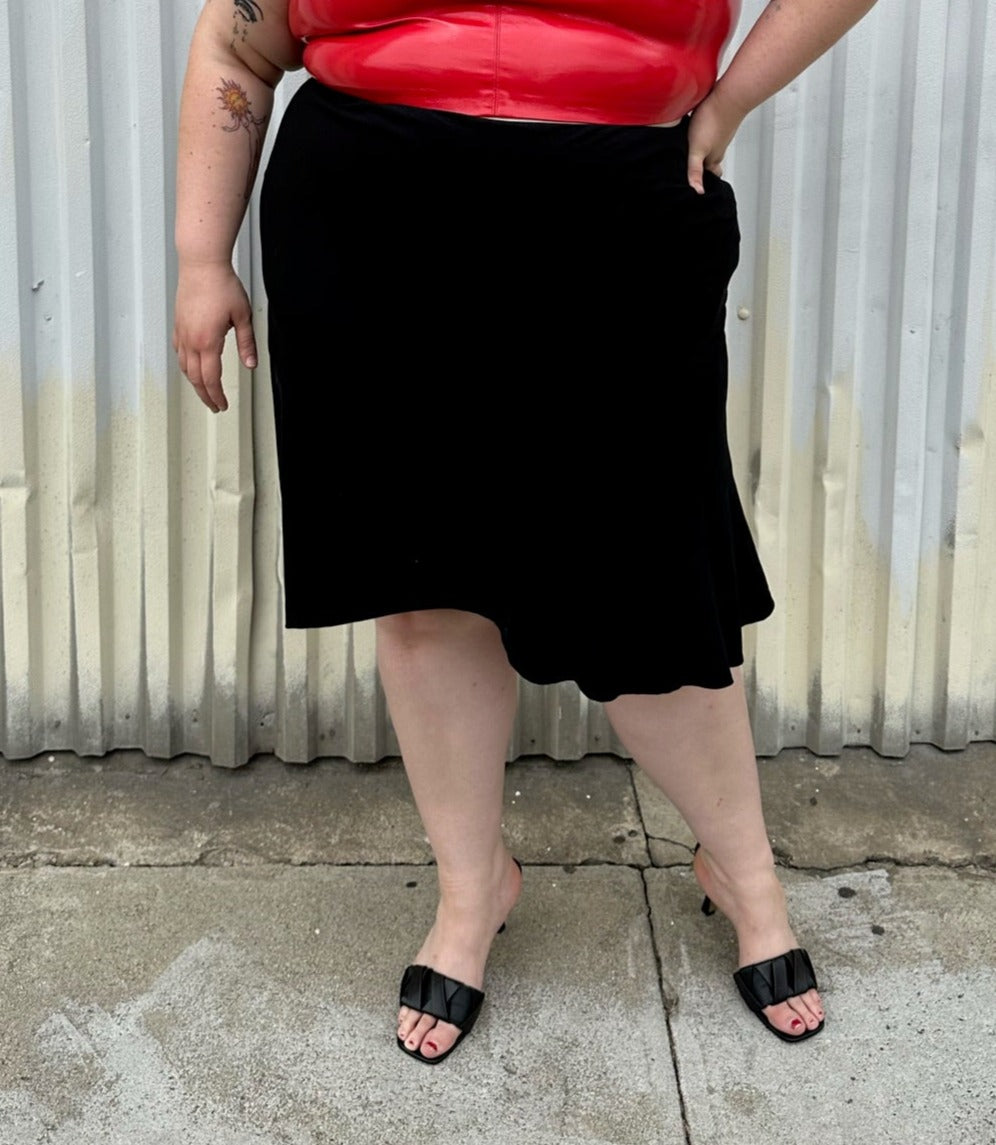 Front view of a size 26 Lane Bryant black handkerchief hem midi skirt with zipper closure styled with a red pleather sleeveless crop top and black kitten heels on a size 22/24 model. The photo is taken outside in natural lighting.