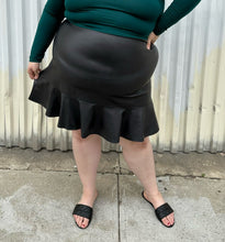 Load image into Gallery viewer, Front view of a size 24 Eloquii black pleather skirt with ruffle hem styled with a teal long sleeve shirt and black slides on a size 22/24 model. The photo is taken outside in natural lighting.

