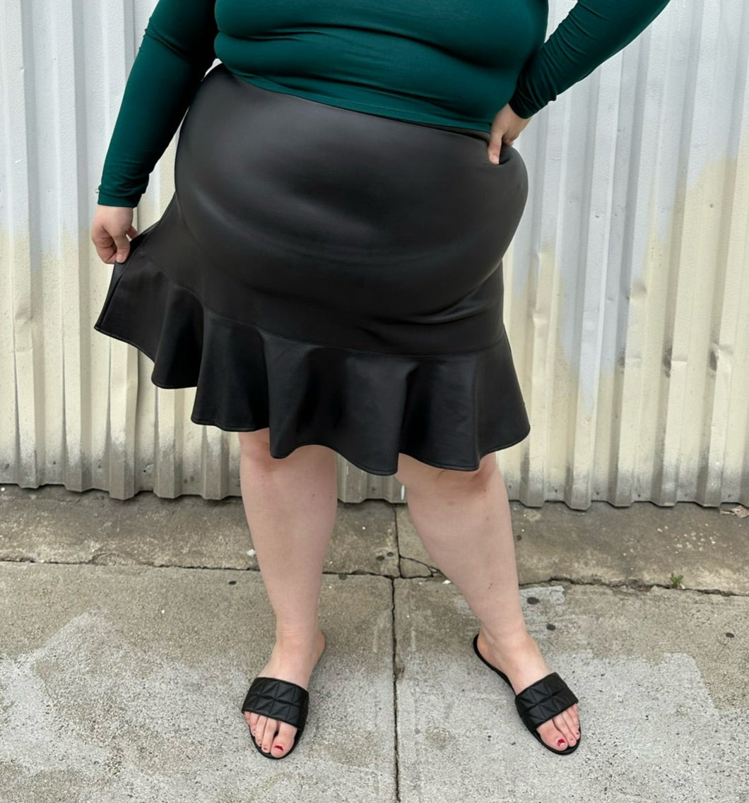 Front view of a size 24 Eloquii black pleather skirt with ruffle hem styled with a teal long sleeve shirt and black slides on a size 22/24 model. The photo is taken outside in natural lighting.