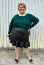 Load image into Gallery viewer, Full-body front view of a size 24 Eloquii black pleather skirt with ruffle hem styled with a teal long sleeve shirt and black slides on a size 22/24 model. The photo is taken outside in natural lighting.
