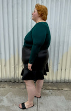 Load image into Gallery viewer, Full-body side view of a size 24 Eloquii black pleather skirt with ruffle hem styled with a teal long sleeve shirt and black slides on a size 22/24 model. The photo is taken outside in natural lighting.
