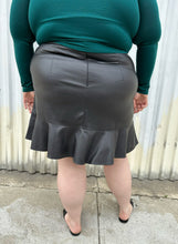 Load image into Gallery viewer, Back view of a size 24 Eloquii black pleather skirt with ruffle hem styled with a teal long sleeve shirt and black slides on a size 22/24 model. The photo is taken outside in natural lighting.
