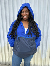 Load image into Gallery viewer, Front view of a size XL Harriton cobalt and navy blue windbreaker styled unzipped with the hood up with jeans on a size 14/16 model. The photo is taken outside in natural lighting.
