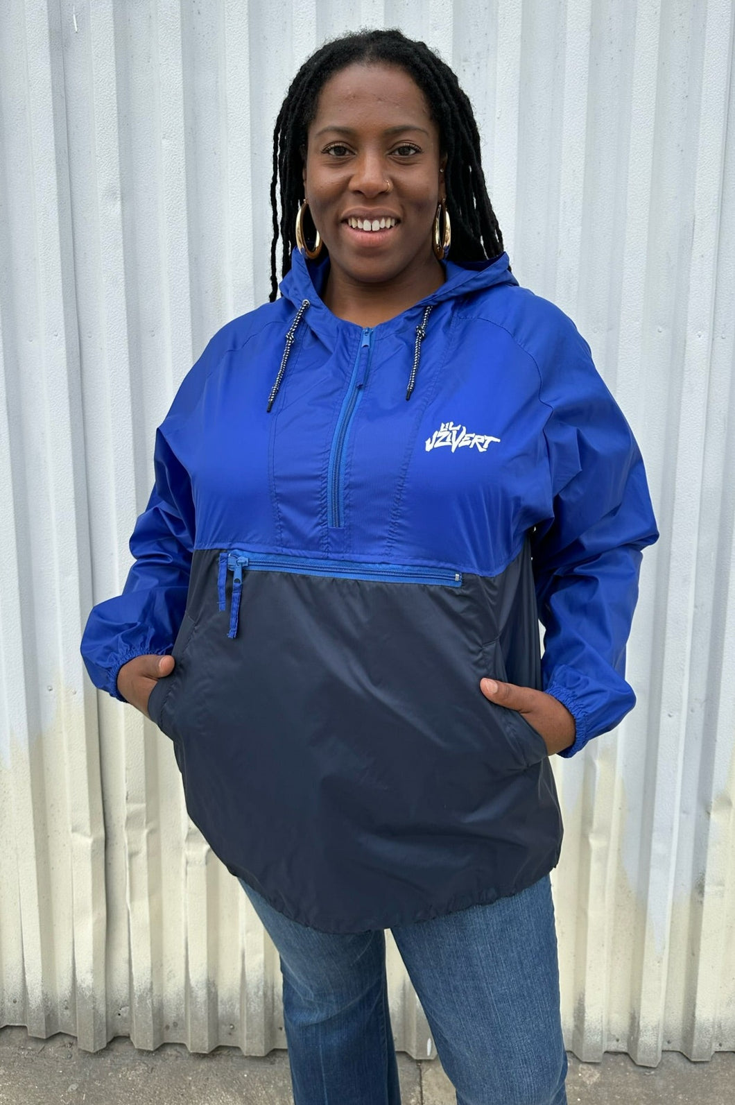 Front view of a size XL Harriton cobalt and navy blue windbreaker styled zipped up with the hood down with jeans on a size 14/16 model. The photo is taken outside in natural lighting.