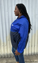 Load image into Gallery viewer, SIde view of a size XL Harriton cobalt and navy blue windbreaker styled zipped up with the hood down with jeans on a size 14/16 model. The photo is taken outside in natural lighting.
