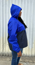 Load image into Gallery viewer, Side view of a size XL Harriton cobalt and navy blue windbreaker styled unzipped with the hood up with jeans on a size 14/16 model. The photo is taken outside in natural lighting.
