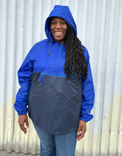 Load image into Gallery viewer, Front view of a size XL Harriton cobalt and navy blue windbreaker styled zipped up with the hood up with jeans on a size 14/16 model. The photo is taken outside in natural lighting.
