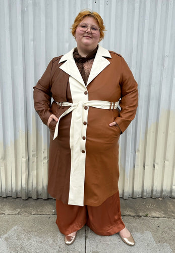 Full-body front view of a size 22/24 Eloquii light brown, dark brown, and cream pleather collared trench coat with contrast colors and belt styled buttoned up and belted over a brown sheer shirt, rust silky pants, and gold flats on a size 22/24 model. The photo is taken outside in natural lighting.