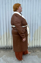 Load image into Gallery viewer, Additional full-body side view of a size 22/24 Eloquii light brown, dark brown, and cream pleather collared trench coat with contrast colors and belt styled buttoned up and belted over a brown sheer shirt, rust silky pants, and gold flats on a size 22/24 model. The photo is taken outside in natural lighting.
