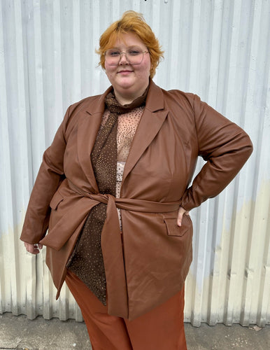 Front view of a size 22 Eloquii warm tone brown belted pleather blazer jacket styled over a dark brown sheer shirt and rust silky pants on a size 22/24 model. The photo is taken outside in natural lighting.