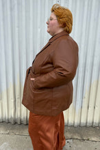 Load image into Gallery viewer, Side view of a size 22 Eloquii warm tone brown belted pleather blazer jacket styled over a dark brown sheer shirt and rust silky pants on a size 22/24 model. The photo is taken outside in natural lighting.
