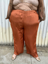 Load image into Gallery viewer, Front view of a pair of size 3X ABLE rust orange colored satin elastic-waist wide leg pants styled with a dark brown sheer shirt and gold flats on a size 22/24 model. The photo is taken outside in natural lighting.
