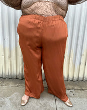 Load image into Gallery viewer, Additional front view of a pair of size 3X ABLE rust orange colored satin elastic-waist wide leg pants styled with a dark brown sheer shirt and gold flats on a size 22/24 model. The photo is taken outside in natural lighting.
