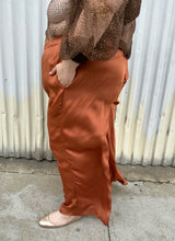 Load image into Gallery viewer, Side view of a pair of size 3X ABLE rust orange colored satin elastic-waist wide leg pants styled with a dark brown sheer shirt and gold flats on a size 22/24 model. The photo is taken outside in natural lighting.
