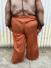 Load image into Gallery viewer, Back view of a pair of size 3X ABLE rust orange colored satin elastic-waist wide leg pants styled with a dark brown sheer shirt and gold flats on a size 22/24 model. The photo is taken outside in natural lighting.
