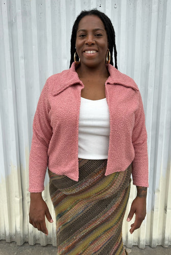 Front view of a LIVD Apparel size 1X dusty rose fuzzy collared crop jacket styled open over a white ribbed tank and earth tone striped skirt on a size 14/16 model. The photo is taken outside in natural lighting.